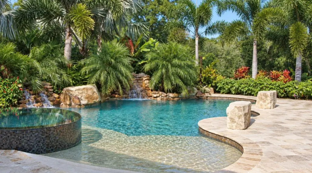 Luxury backyard swimming pool with turquoise water, a raised circular spa, natural stone decking, tropical palm trees, and cascading rock waterfalls.