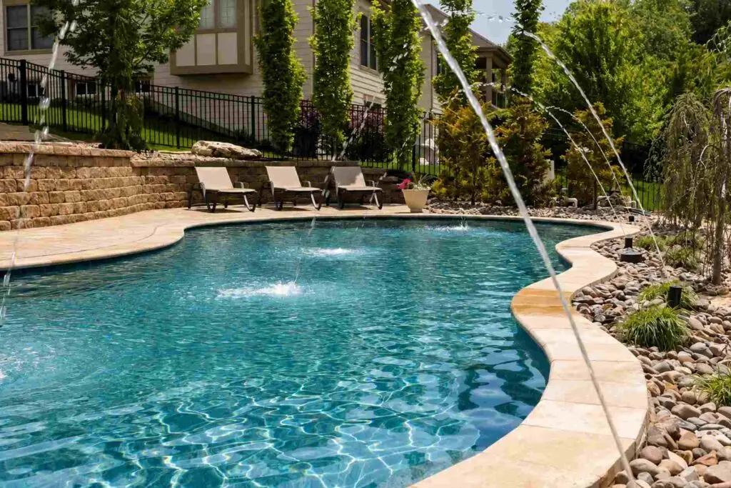 Freeform backyard pool with arcing deck jets, cream stone coping and a tiered block retaining wall lined with lounge chairs