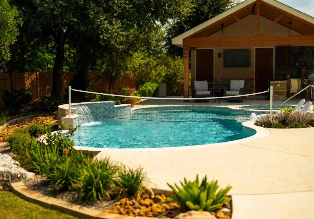 Freeform pool with a volleyball net and tiled waterfall wall, facing a covered cabana with an outdoor kitchen