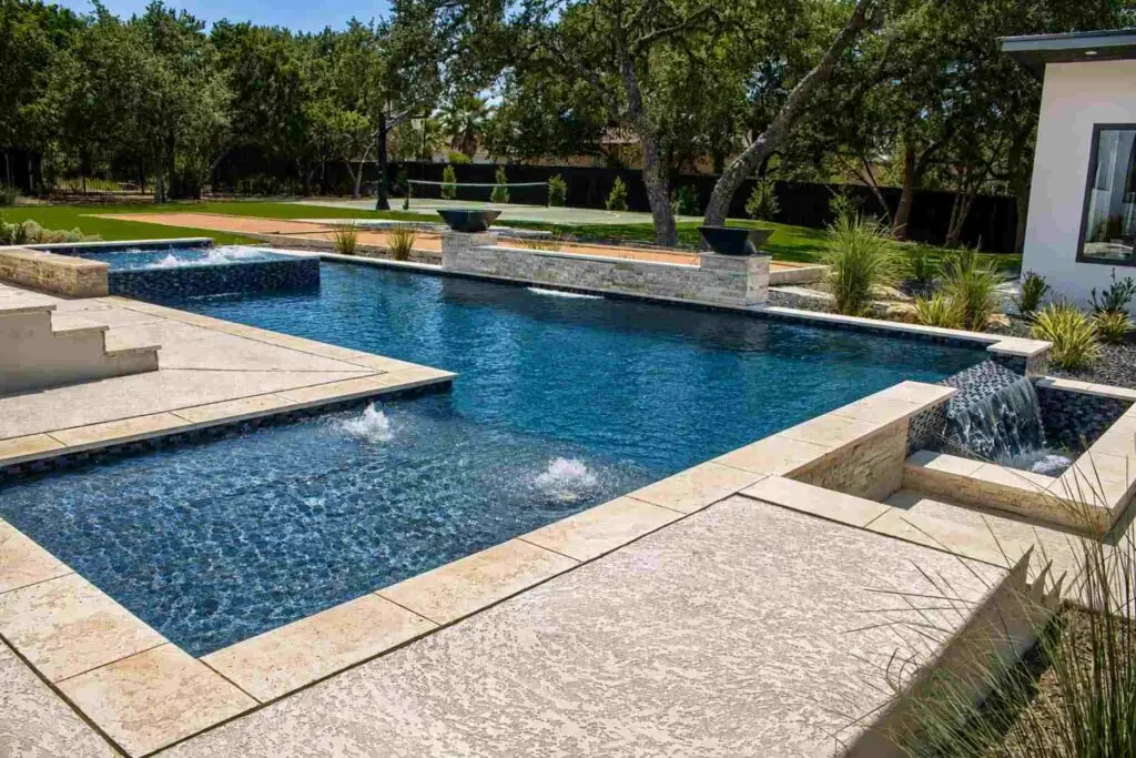 Modern rectangular pool with raised spa, bubblers, dark glass tile spillways and black fire bowls on a travertine deck