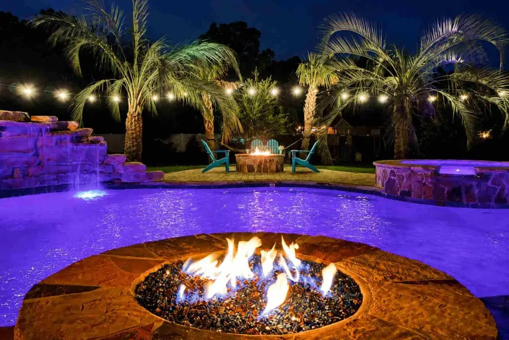 Backyard pool glowing purple at night with a lit fire pit, rock waterfall, spa and string-lit palm trees
