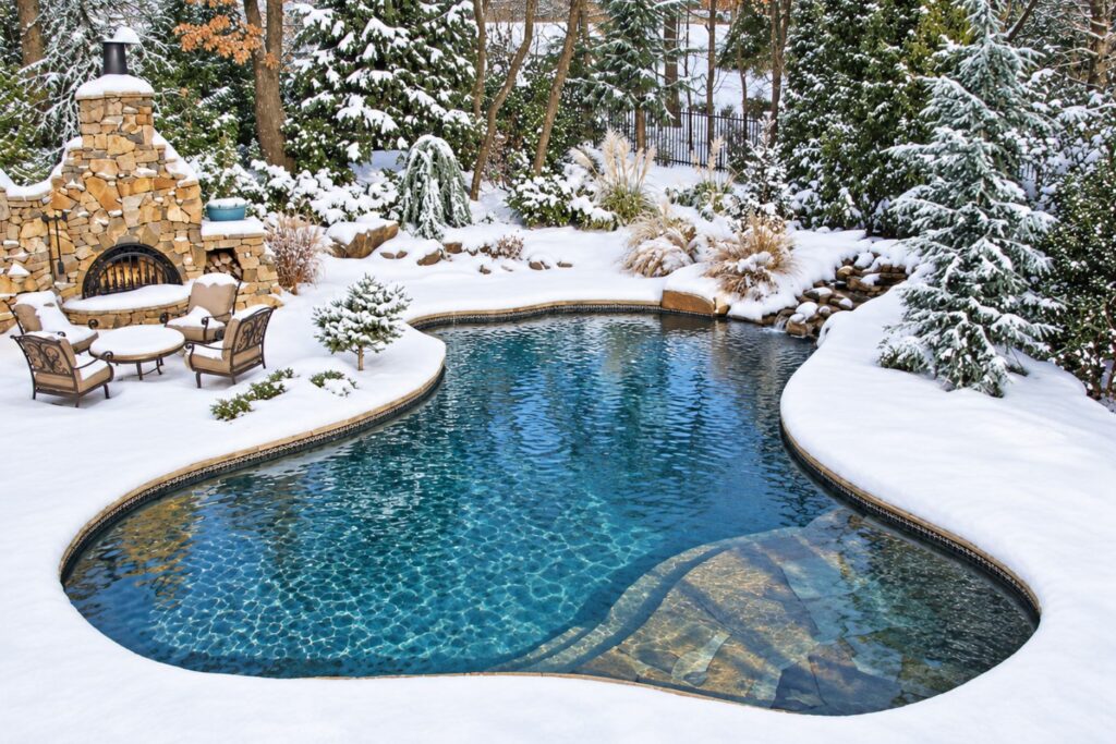 Freeform inground pool surrounded by snow with a stone outdoor fireplace and patio chairs
