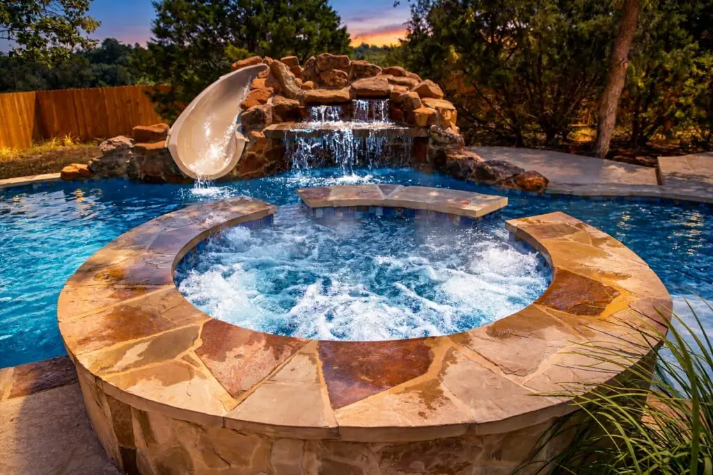 Bubbling raised stone hot tub with a rock waterfall and pool slide behind it at dusk