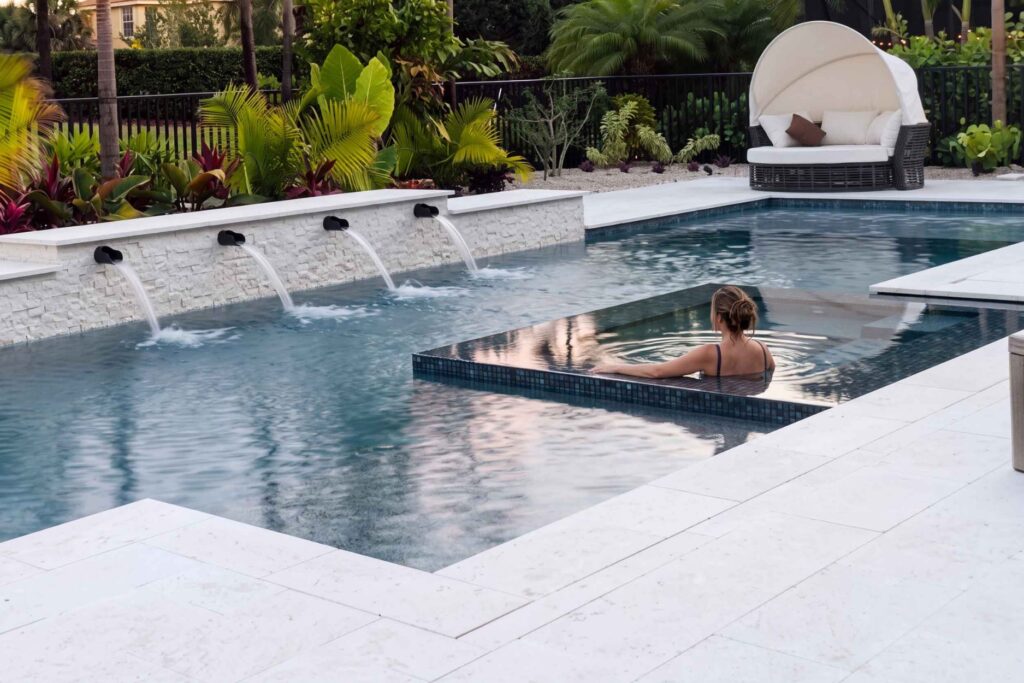 Modern pool and spa combo with a raised spillover spa, sheer descent water features, and white stone decking in a tropical backyard