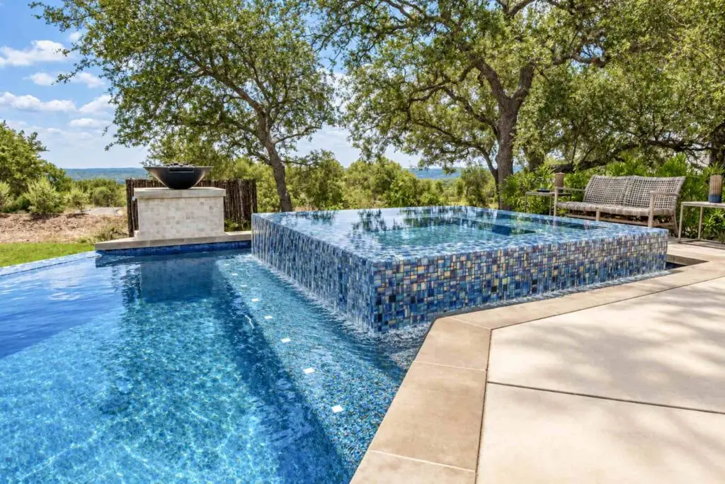 Raised spa with blue glass tile and water spilling over every edge into a backyard swimming pool