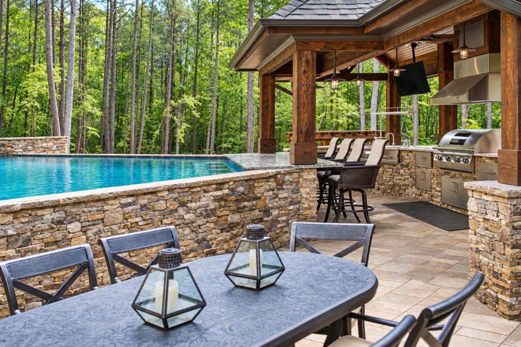 Poolside outdoor kitchen under a timber pavilion with granite bar seating, grill, and a dining table beside the pool