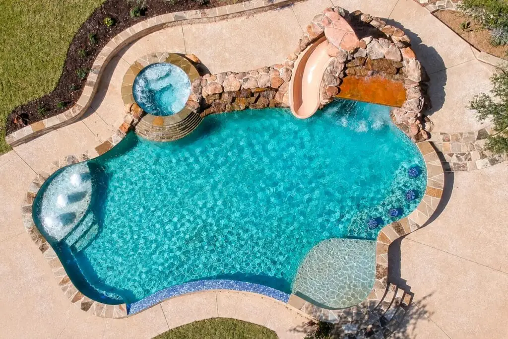 Aerial view of a freeform pool with a tan waterslide, rock waterfall wall, raised spa, tanning ledge and stamped concrete deck