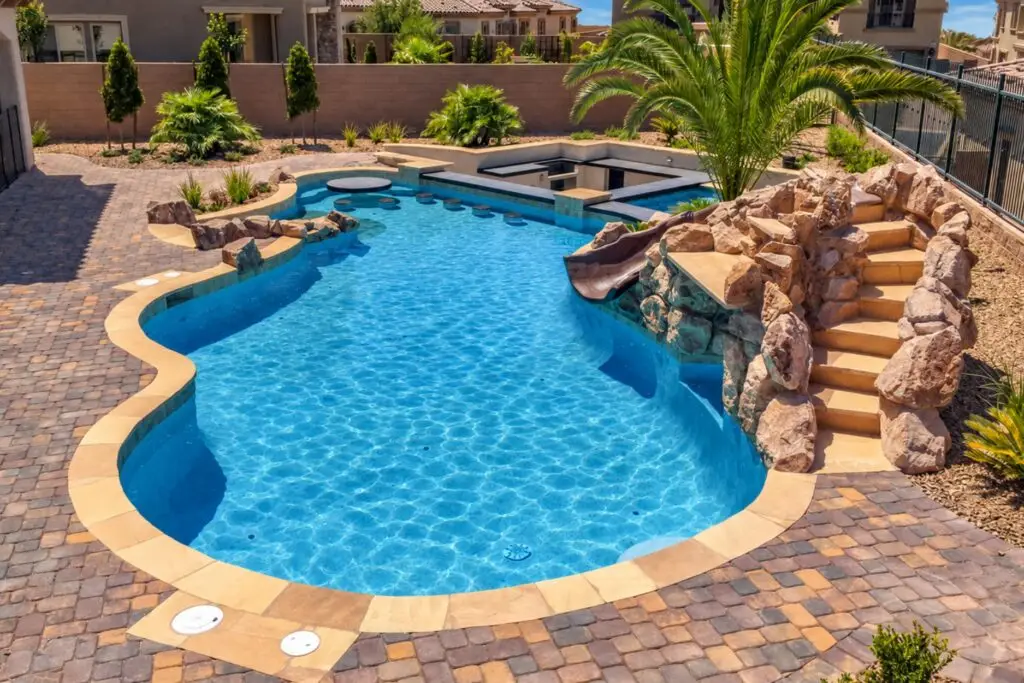 Tan flagstone pool coping framing a freeform pool with a rock slide, swim-up bar, and brick paver deck
