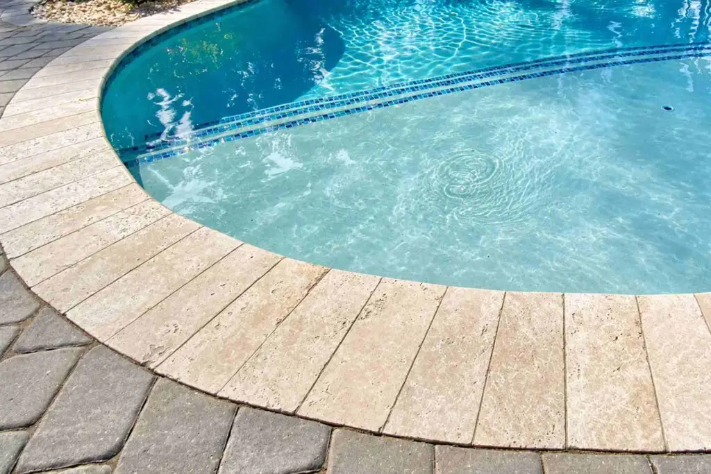 Travertine pool coping on a curved pool edge next to a gray paver deck