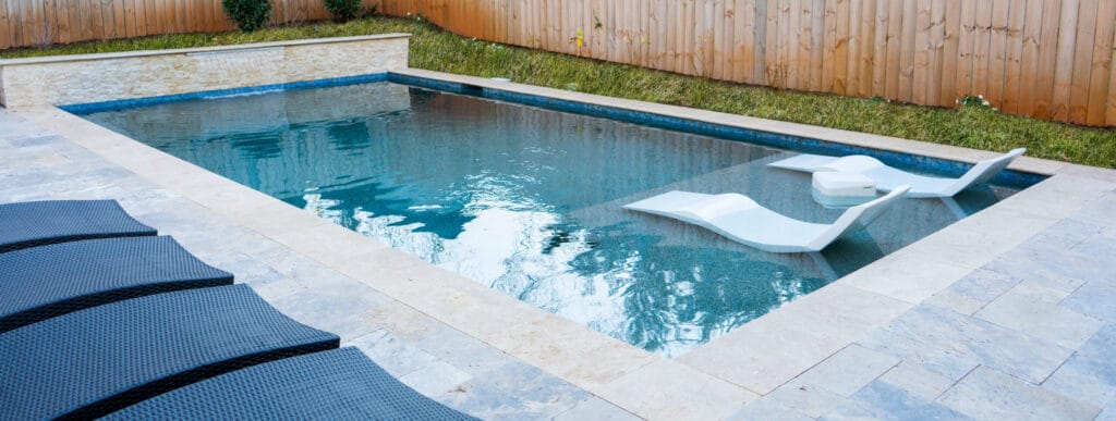 Rectangular pool with tanning ledge and chaise lounges in fenced backyard.
