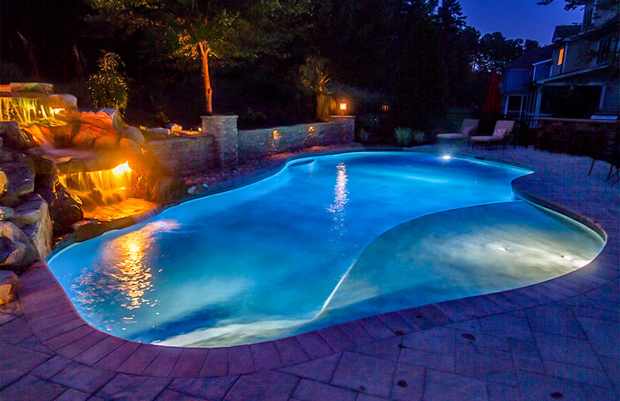 Freeform custom pool with blue LED lighting at night, featuring a rock grotto waterfall with warm accent lights and stamped concrete patio