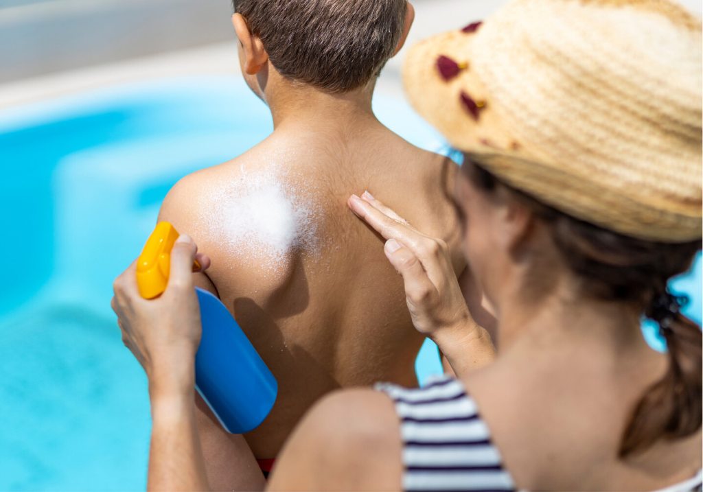 Mother in a straw sun hat applying sunscreen to her young son's back beside a backyard swimming pool