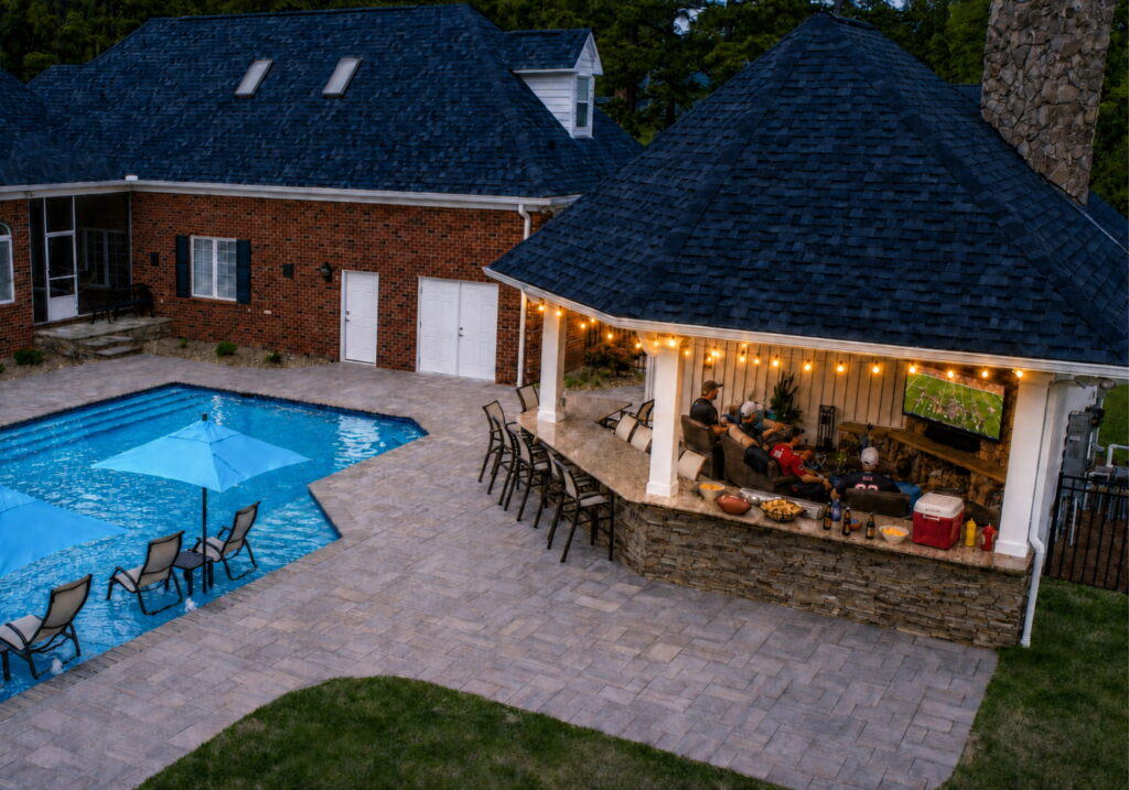 Guests watching football on an outdoor TV under a lit poolside cabana at dusk, with snacks and a cooler on the stone bar and a paver patio around the pool.