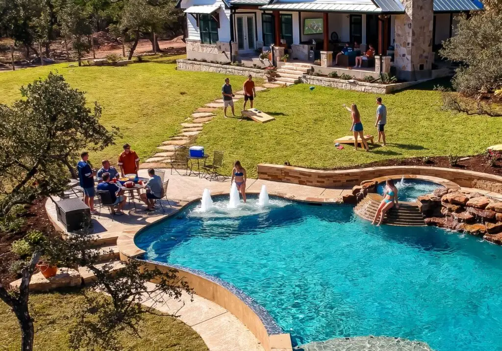 Aerial view of a backyard pool party with bubbling deck jets, a raised spa with rock waterfall, patio dining, and cornhole on the lawn
