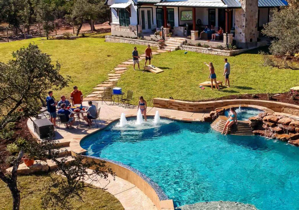 Aerial view of a backyard pool party with bubbling deck jets, a raised spa with rock waterfall, patio dining, and cornhole on the lawn as a backyard tailgating game