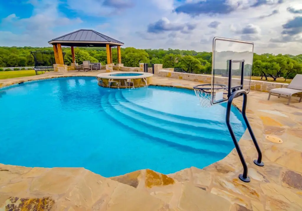 Freeform gunite pool with poolside basketball hoop backyard tailgating game, raised round spa with spillways, and flagstone deck overlooking Hill Country views