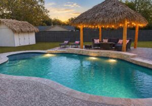 Freeform pool at sunset with lit sheer descent waterfalls and a thatched palapa with string lights over patio seating