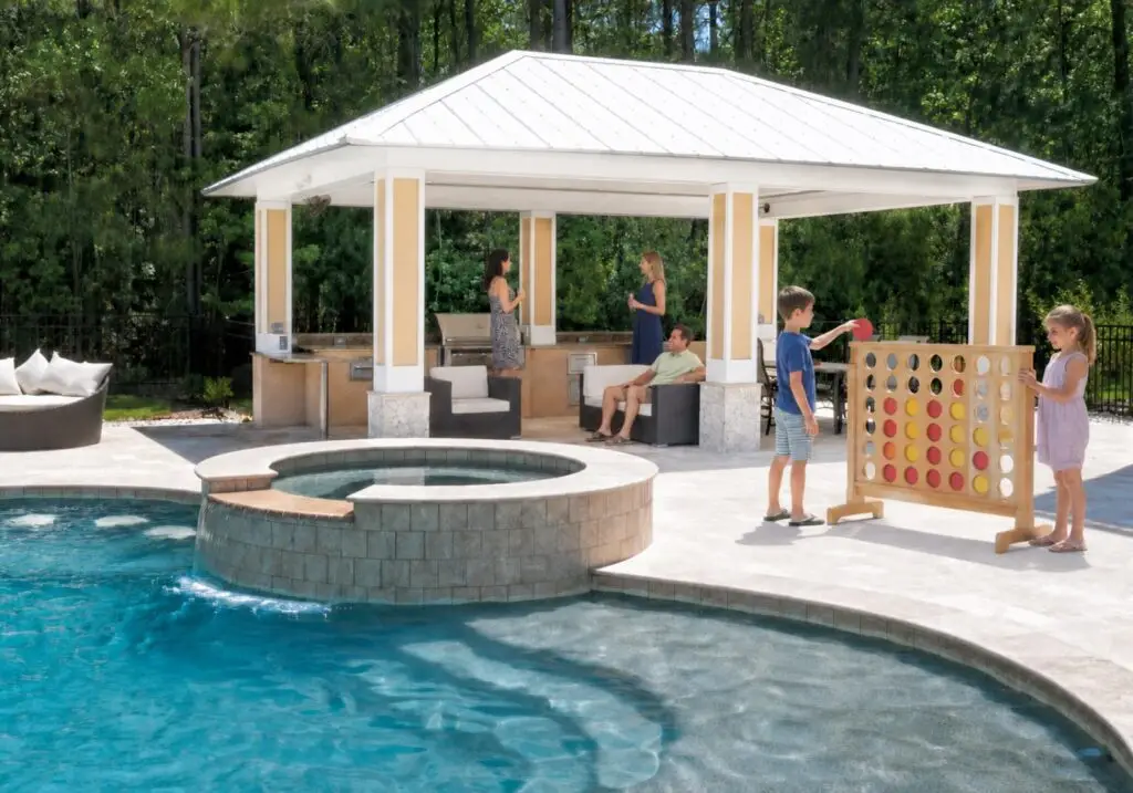 Family enjoying a poolside pavilion with outdoor kitchen and lounge seating beside a pool with a raised spa spillover and giant Connect Four game as a backyard tailgating game