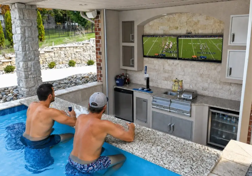 Two men in a swim-up pool bar watching football on two TVs in a covered outdoor kitchen.