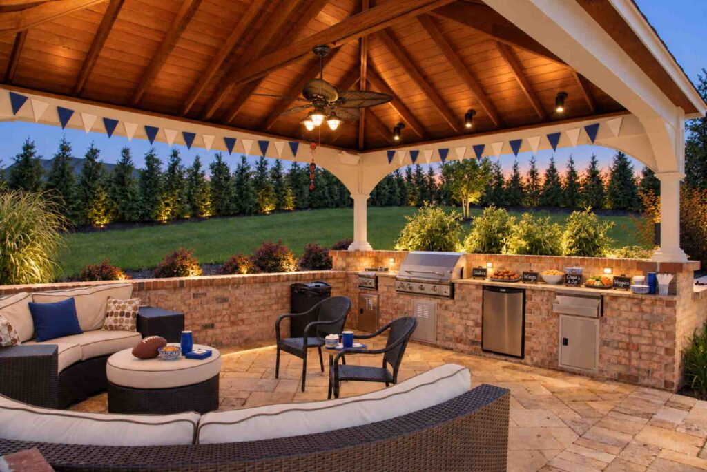 Game-day party in a covered pavilion with a brick outdoor kitchen, built-in grill, snack spread, and wicker seating at dusk.