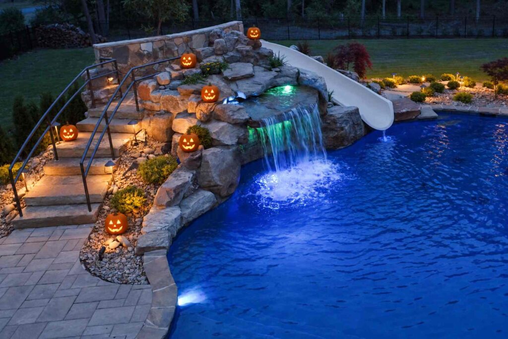 Backyard pool at night decorated for Halloween, with jack-o'-lanterns along stone steps and a rock waterfall, a lit waterfall pouring into blue water, and a white water slide.