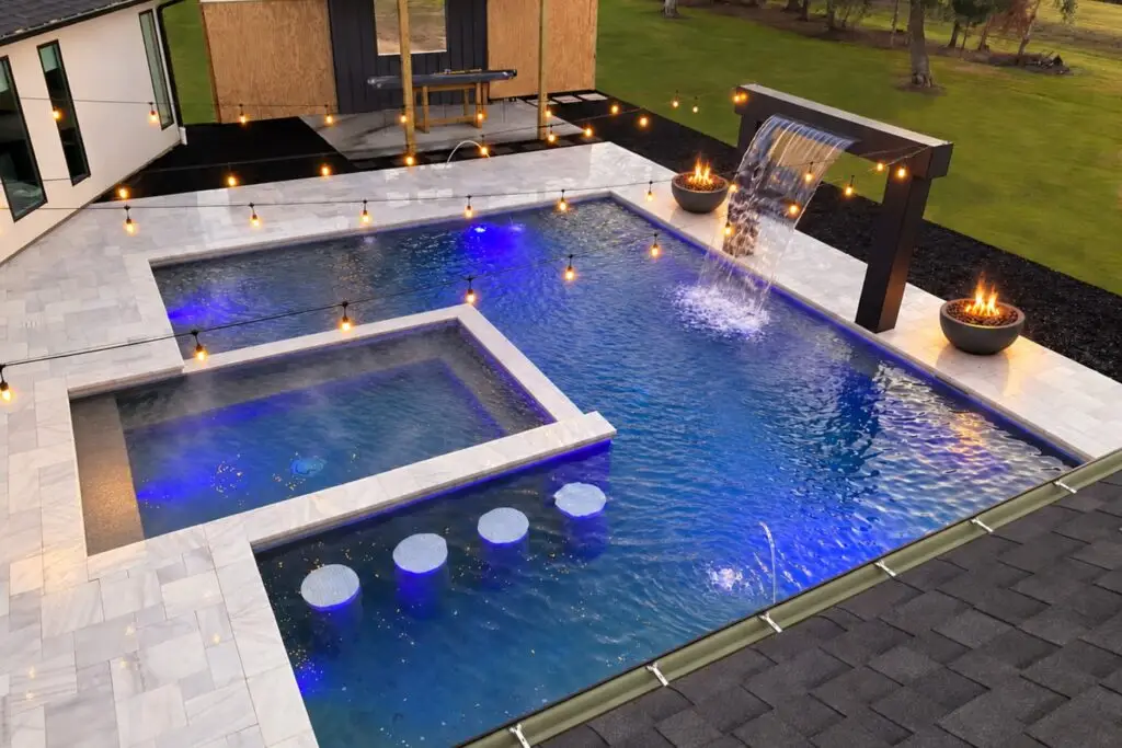 Aerial view of a modern rectangular pool and raised spa at dusk with blue LED lighting, swim-up bar stools, a sheer descent waterfall, fire bowls and string lights over a white paver deck.