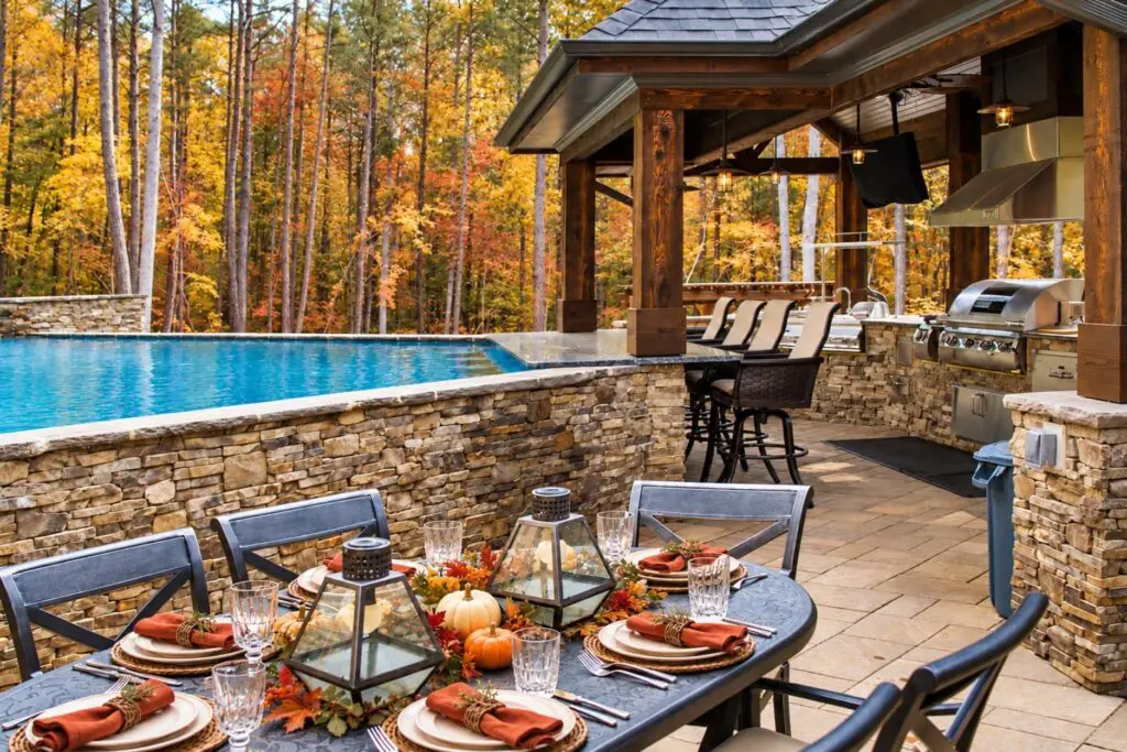 Fall table set with pumpkins and lanterns beside a raised pool with a stone wall, a swim-up bar, and a covered outdoor kitchen with a built-in grill, surrounded by autumn trees.