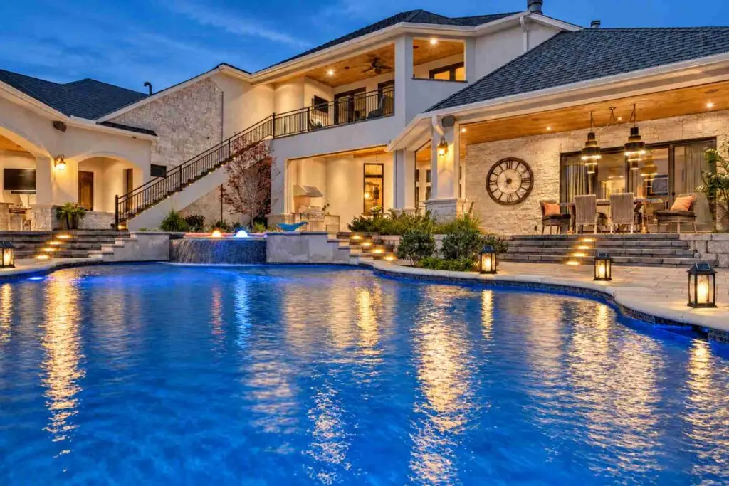 Freeform pool glowing blue at dusk, with a sheer descent waterfall, lit stone steps, and lanterns along the deck in front of a two-story home with a covered patio.