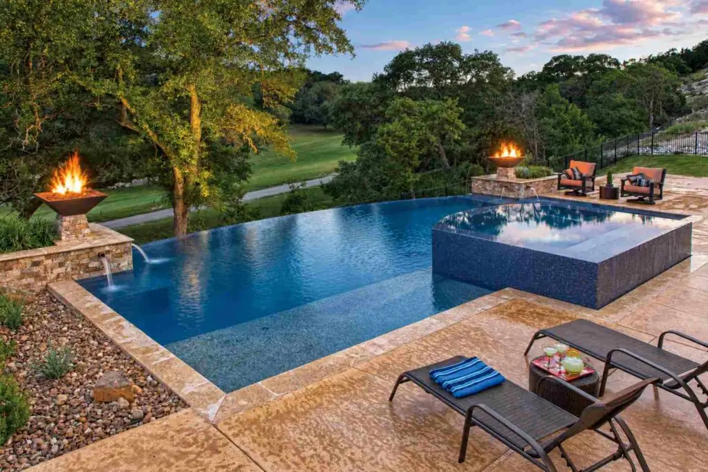Infinity edge pool with a raised tiled spa, fire bowls, and scupper water features, overlooking a green hillside, with lounge chairs and drinks on a stone deck.